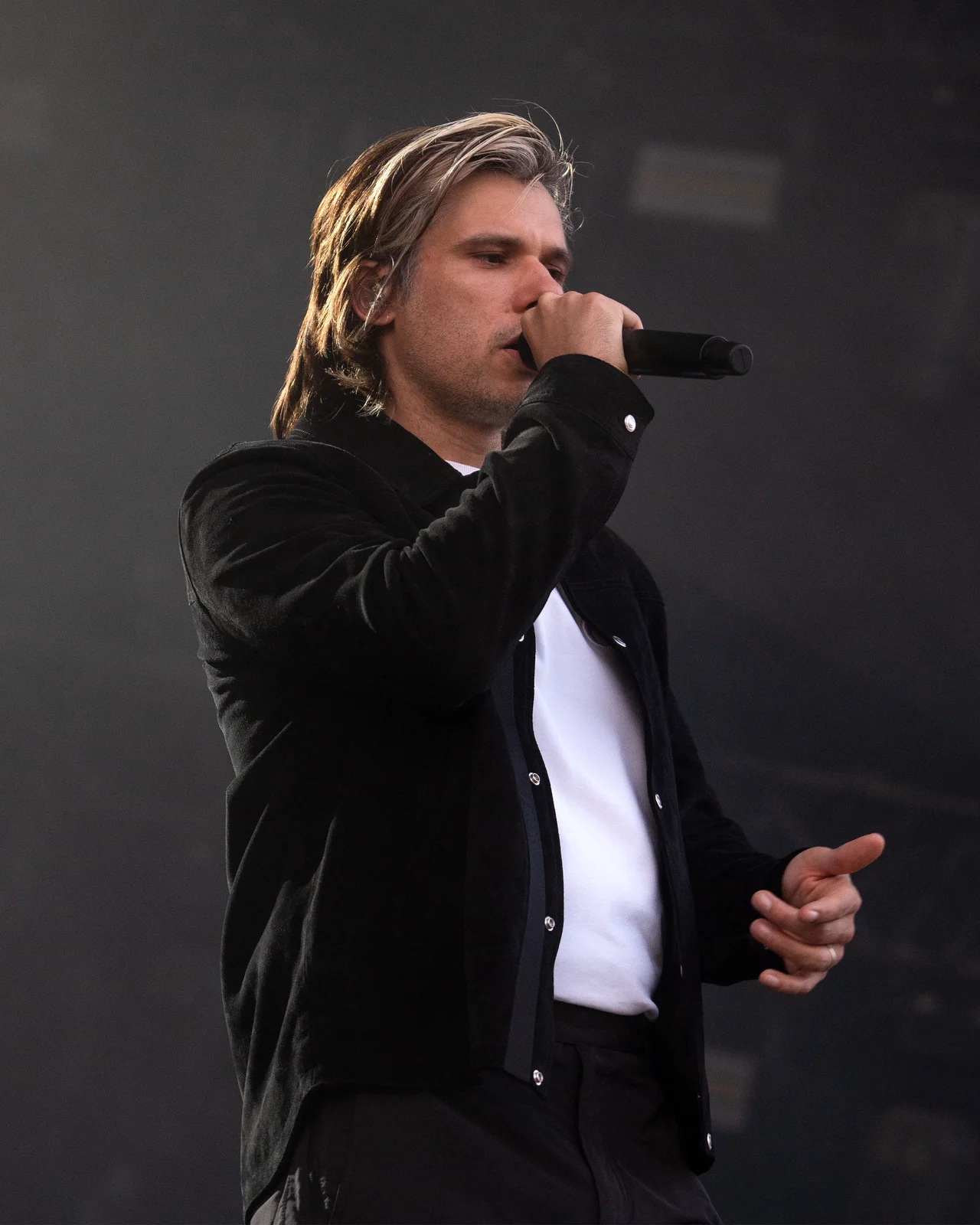 Orelsan en veste noire et tee-shirt blanc, chantant au micro sur fond sombre.