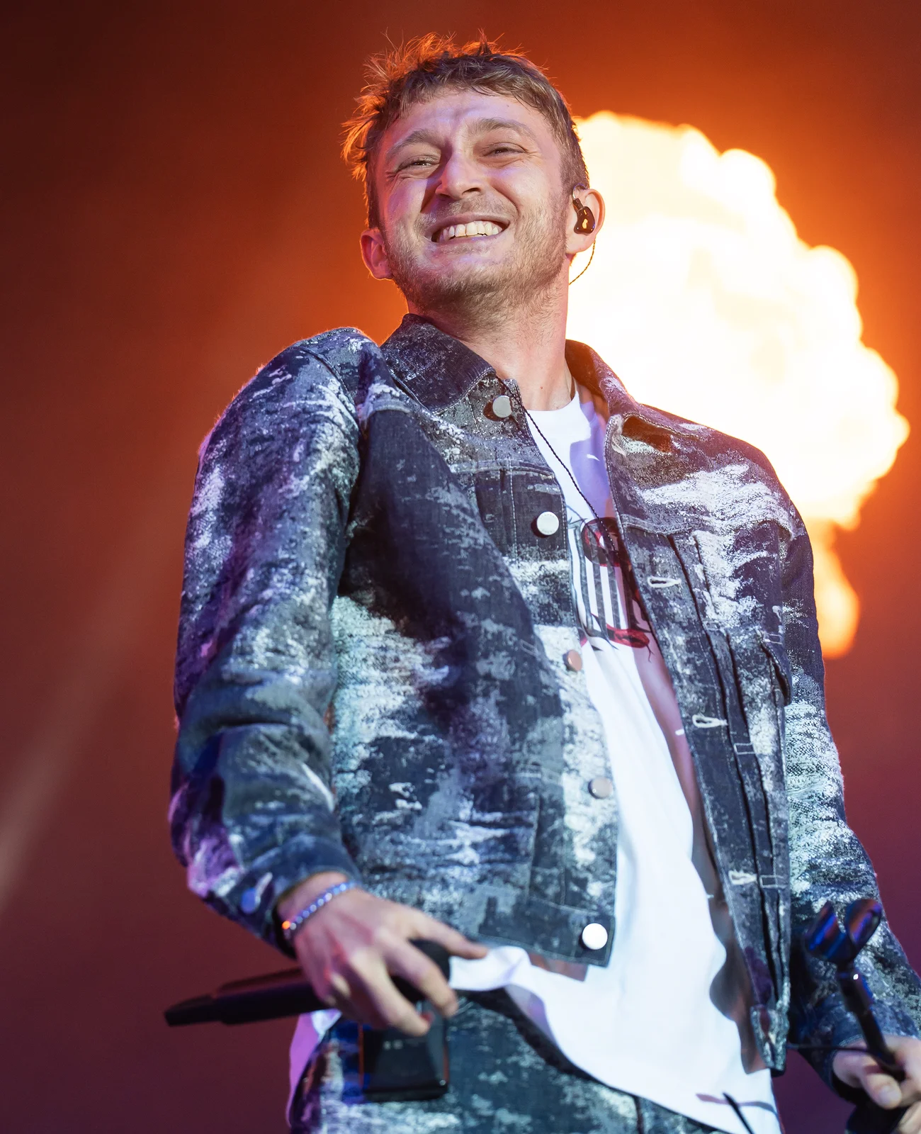 Vald tout sourire en veste en jean délavé, devant une gerbe de flammes sur scène.