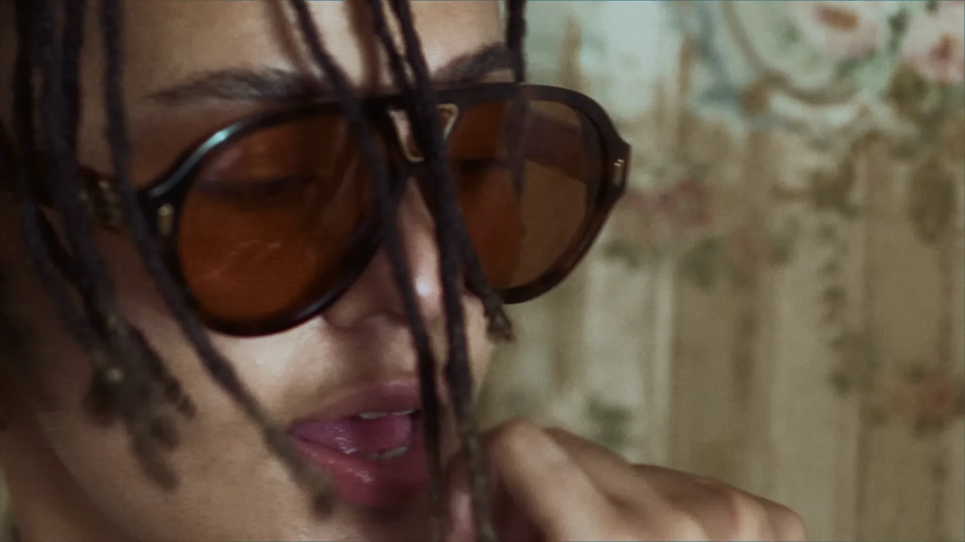 Gros plan sur Lala &ce, locks sur le visage et grandes lunettes aux verres orangés, devant un papier peint fleuri.