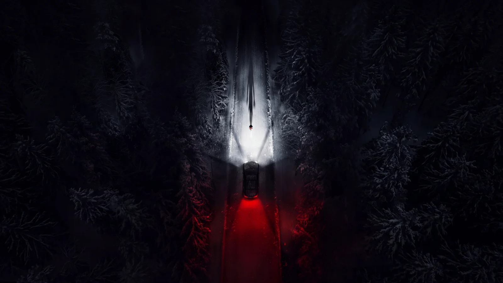 Vue aérienne d’une route enneigée traversant une forêt de nuit, une voiture éclairée de rouge et une silhouette dans la lumière des phares, dans Resident Evil.