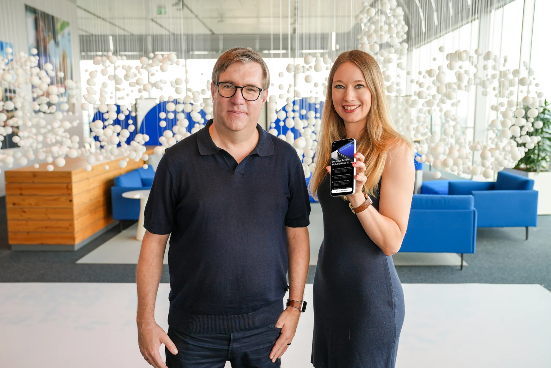 Un homme à lunettes en polo bleu marine et une femme en robe grise, qui tient un smartphone affichant une application, posent dans un hall de bureaux décoré de sphères blanches suspendues et de canapés bleus.