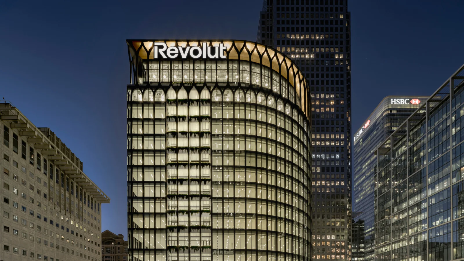 Le siège de Revolut à Canary Wharf, à Londres, photographié de nuit : une tour de verre éclairée, coiffée de l’enseigne Revolut, entre d’autres immeubles de bureaux, dont la tour HSBC à droite.