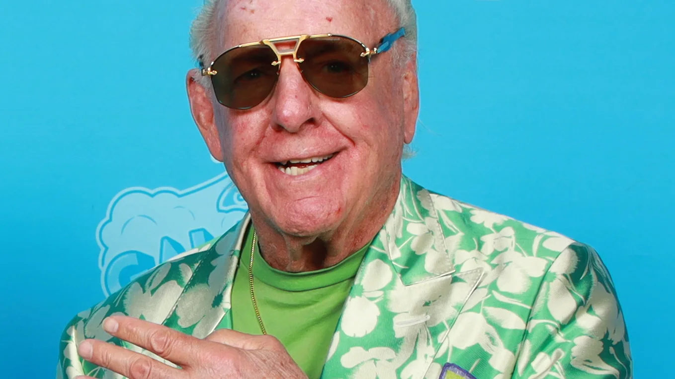 Ric Flair, lunettes de soleil dorées et veste à motifs floraux verts, sourit devant un fond bleu.