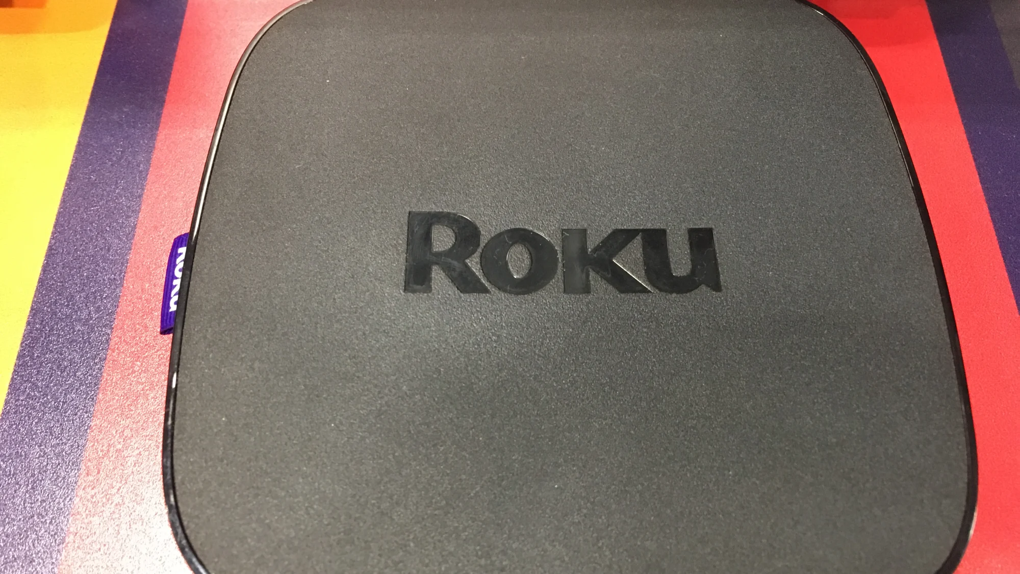 Un boîtier de streaming Roku Ultra noir, le logo Roku gravé sur le dessus, posé sur un présentoir rouge et violet.