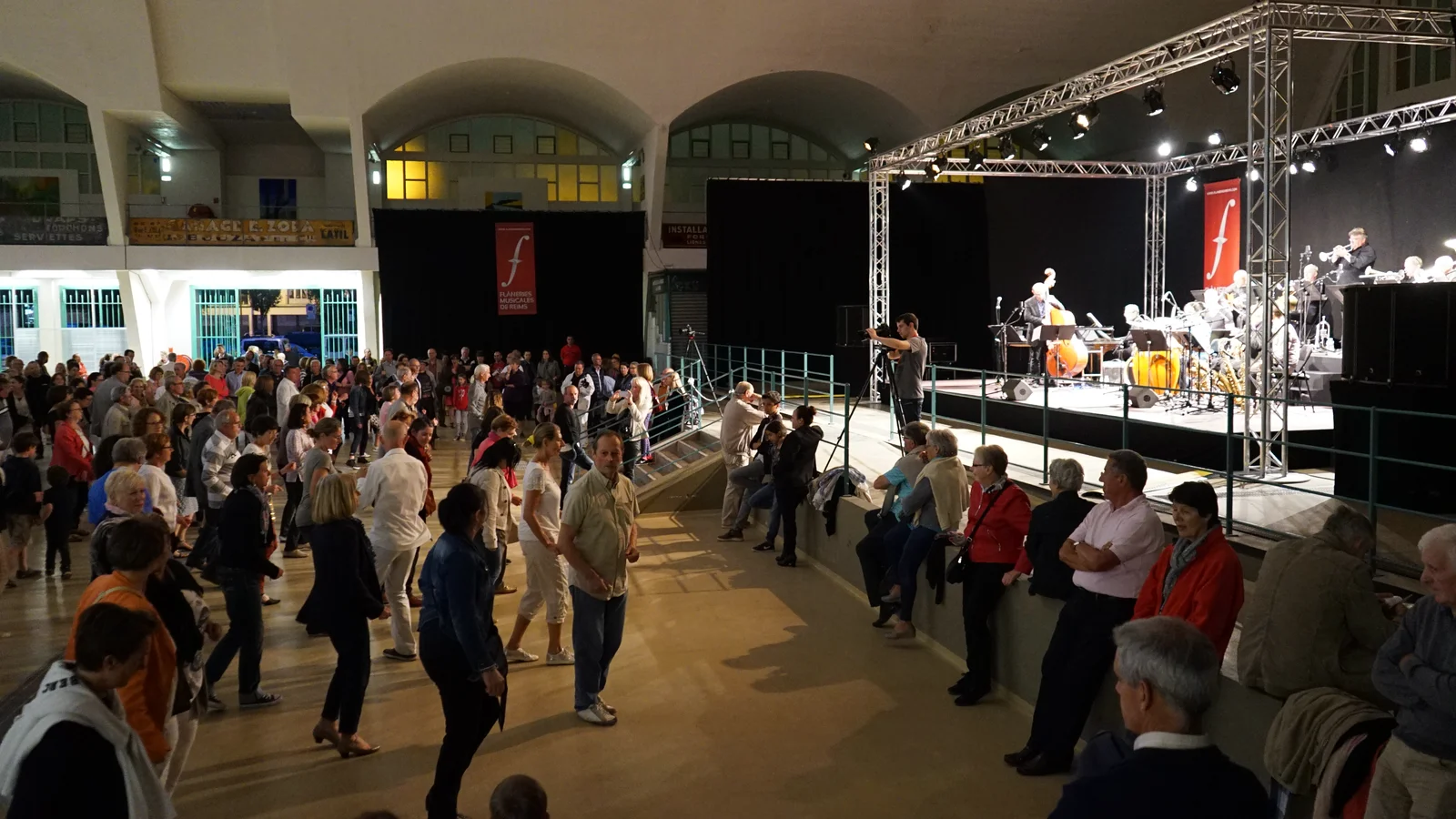Un bal populaire dans une grande halle : des couples dansent sur le parquet devant une scène où joue un orchestre de jazz, des spectateurs accoudés à la rambarde.