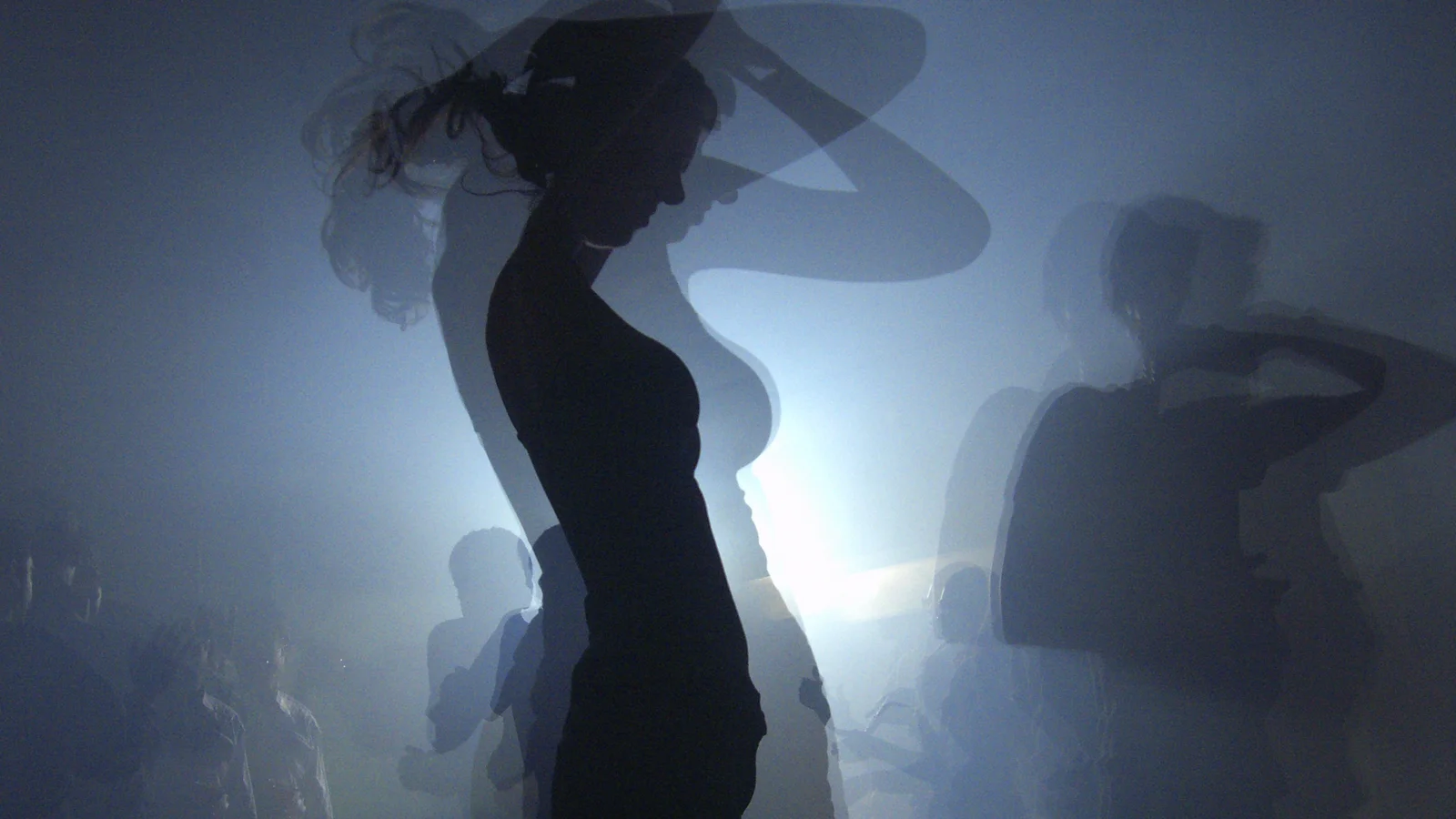 Silhouettes de danseurs en contre-jour sur une piste noyée de fumée bleutée, au premier plan une danseuse de profil, les bras levés.