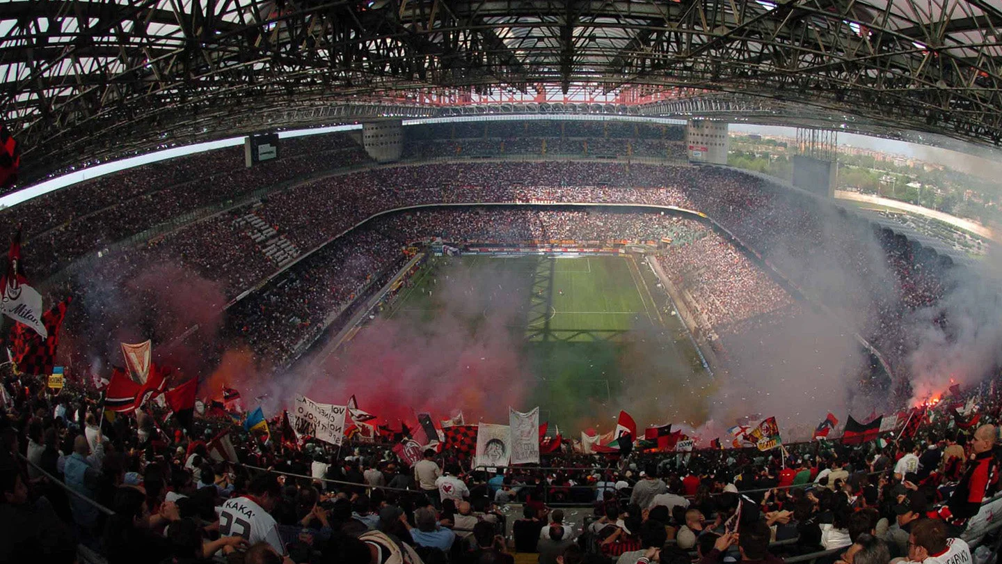 San Siro vu de haut depuis la tribune rossonera, sous les poutres du toit : les gradins pleins, la pelouse au centre, des drapeaux rouge et noir et des nuées de fumigènes qui montent dans la lumière du jour.