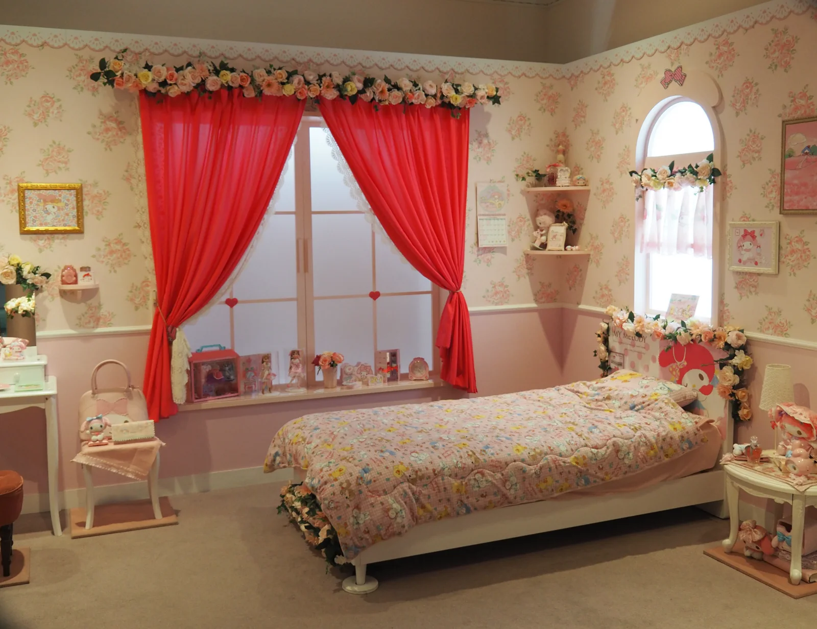 Une chambre d’enfant rose reconstituée, avec rideaux rouges, guirlandes de roses, lit fleuri et peluches, décorée aux couleurs du personnage My Melody.