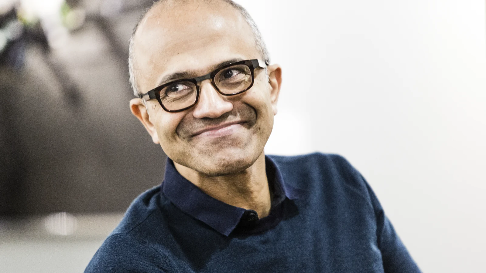 Satya Nadella, PDG de Microsoft, souriant, en pull bleu marine à col polo, sur un fond clair.