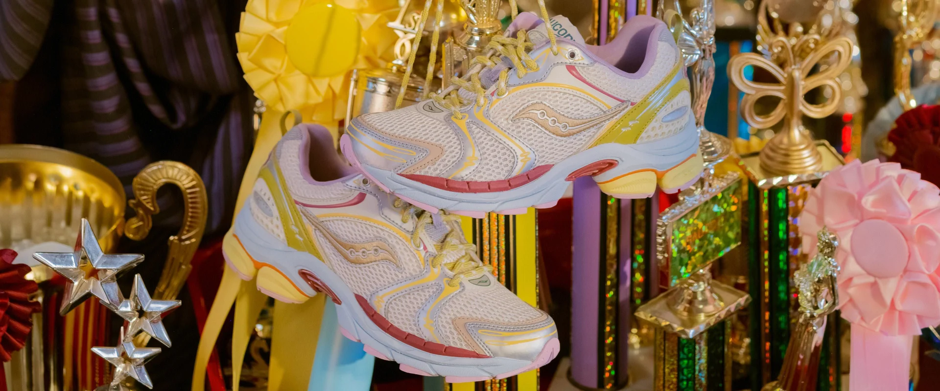 La paire suspendue par ses lacets devant une vitrine remplie de trophées dorés, d’étoiles argentées et de rosettes jaune, rose et rouge.