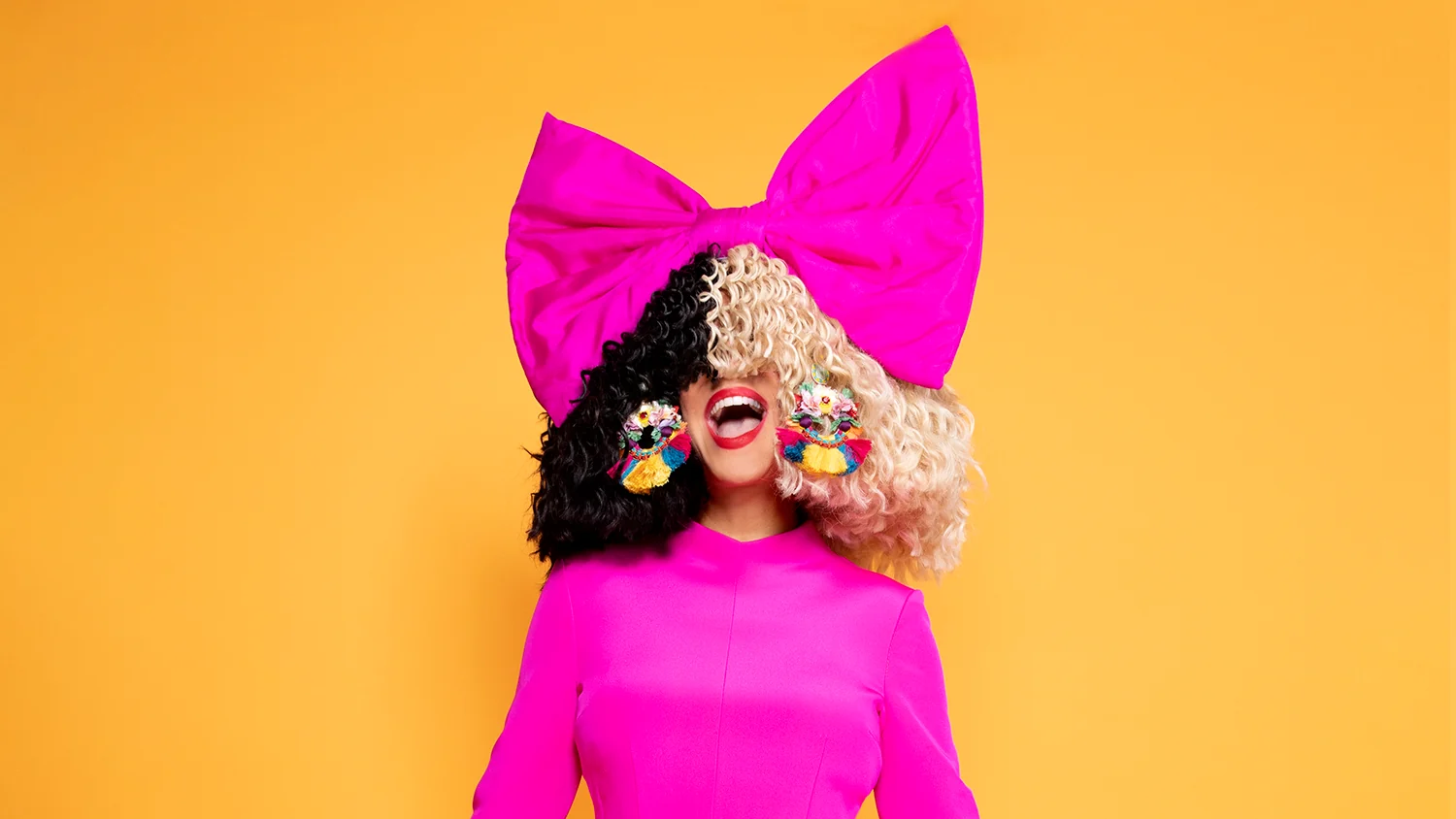 Portrait en studio de Sia : un grand nœud rose fuchsia sur une perruque mi-noire mi-blonde qui cache ses yeux, bouche rouge ouverte en un grand rire, boucles d’oreilles multicolores, robe fuchsia, sur fond orange.