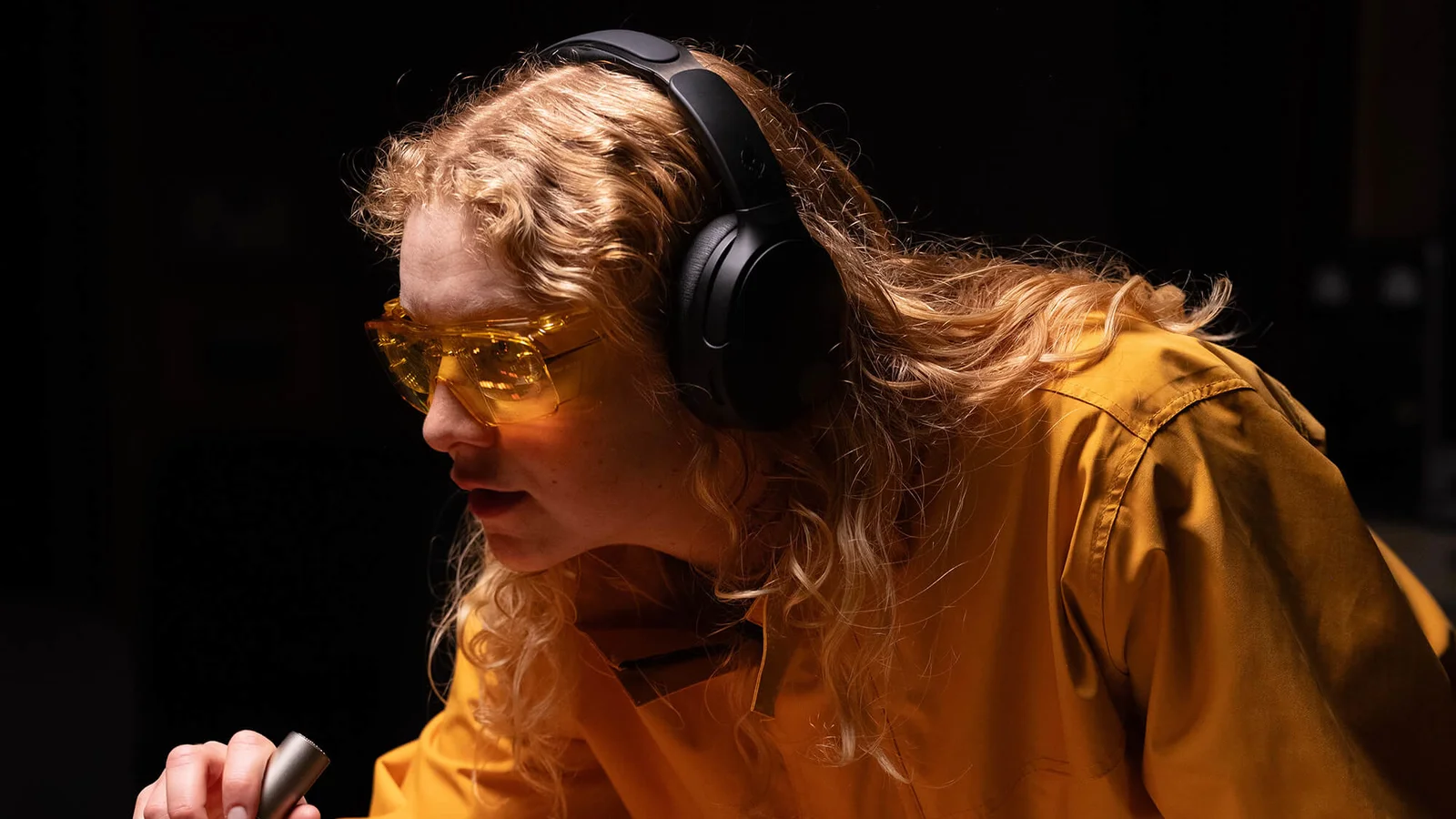 Une jeune femme aux cheveux bouclés, lunettes ambrées et veste orange, porte un casque noir Skullcandy Crusher 1080 ANC et se penche vers une console de studio.