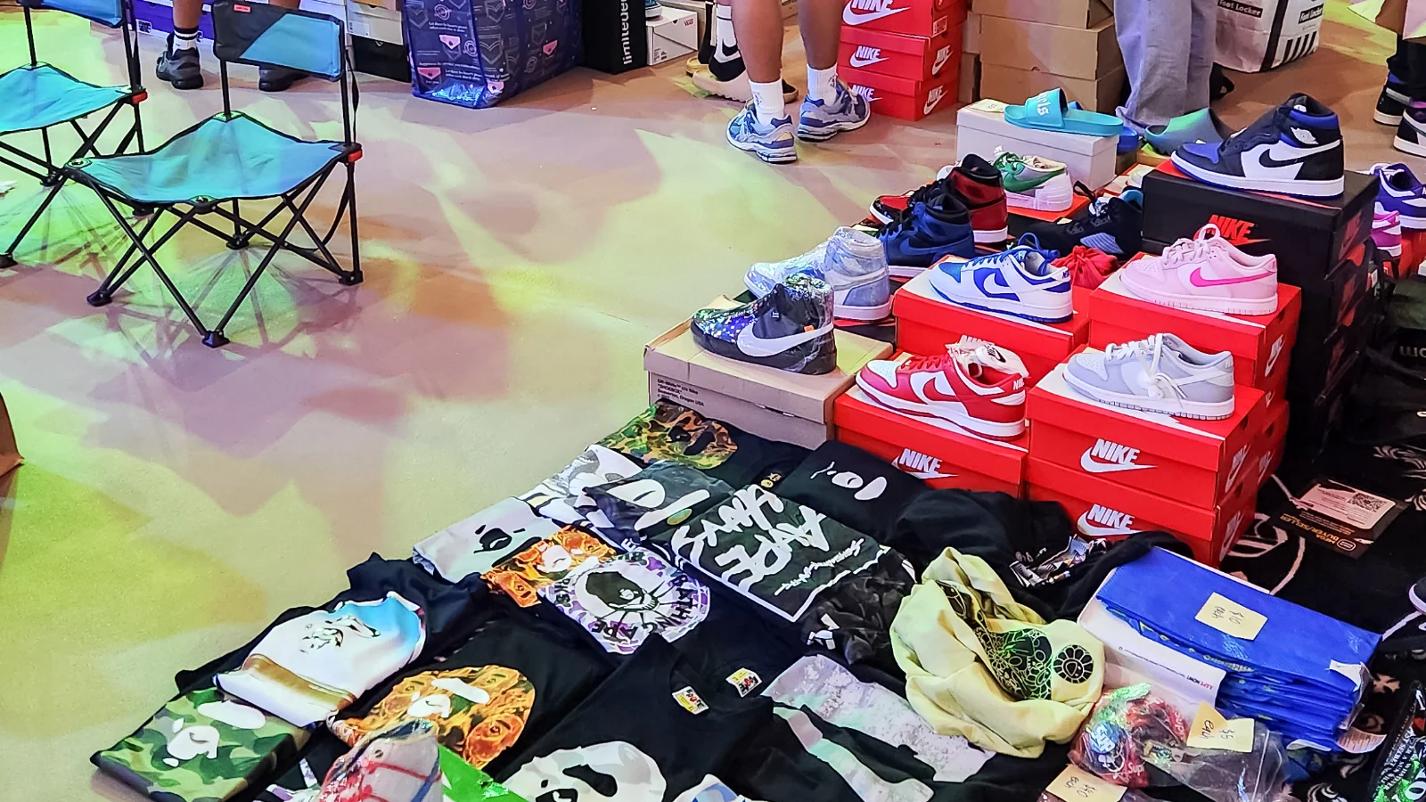Allée d'un salon de sneakers : des dizaines de paires et de boîtes orange posées au sol, des visiteurs qui circulent entre les stands sous un plafond orné de fleurs jaunes et rouges.