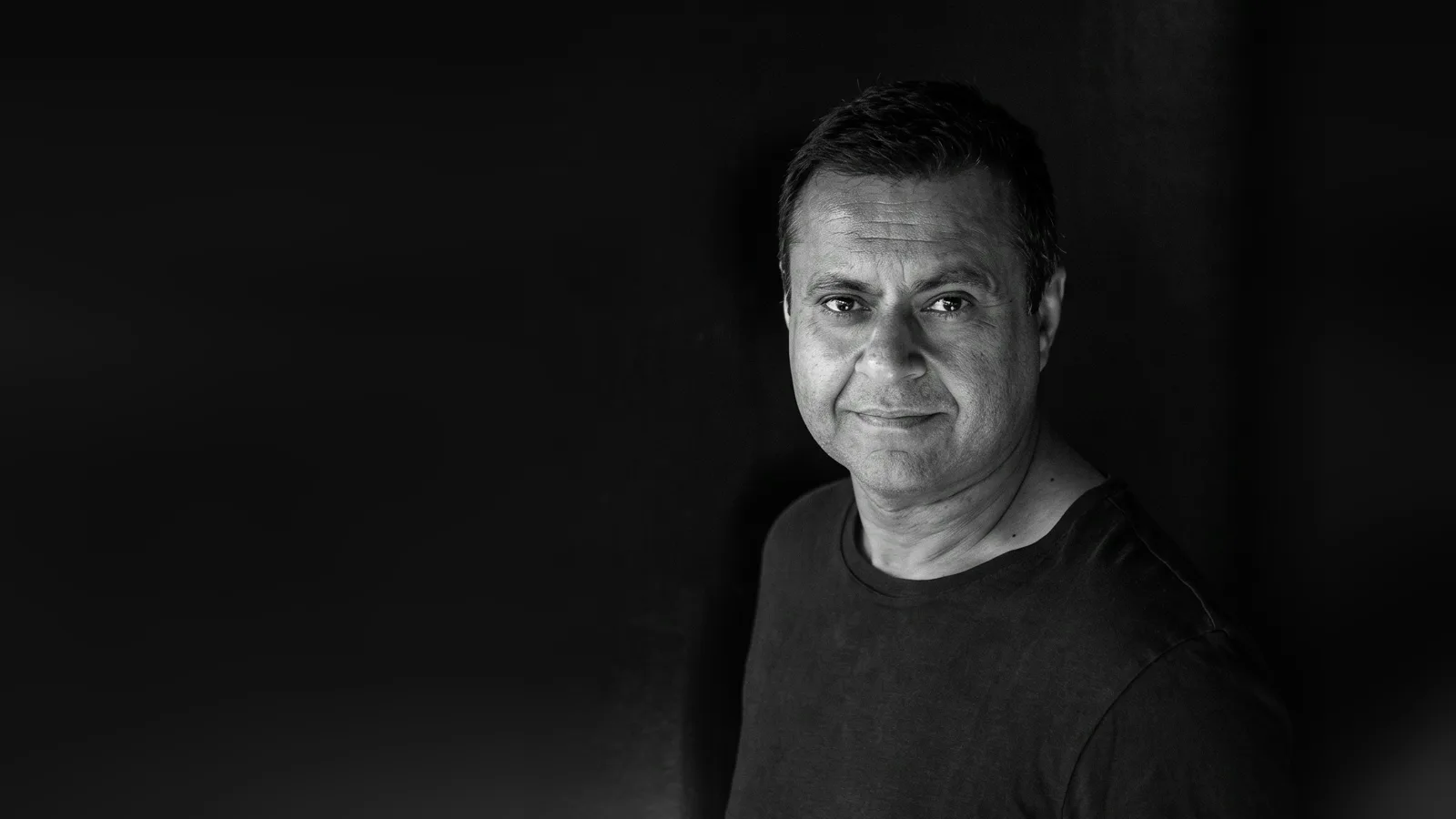 Portrait en noir et blanc de Sonny Takhar, en tee-shirt sombre, qui sourit légèrement face à l’objectif sur un fond noir.