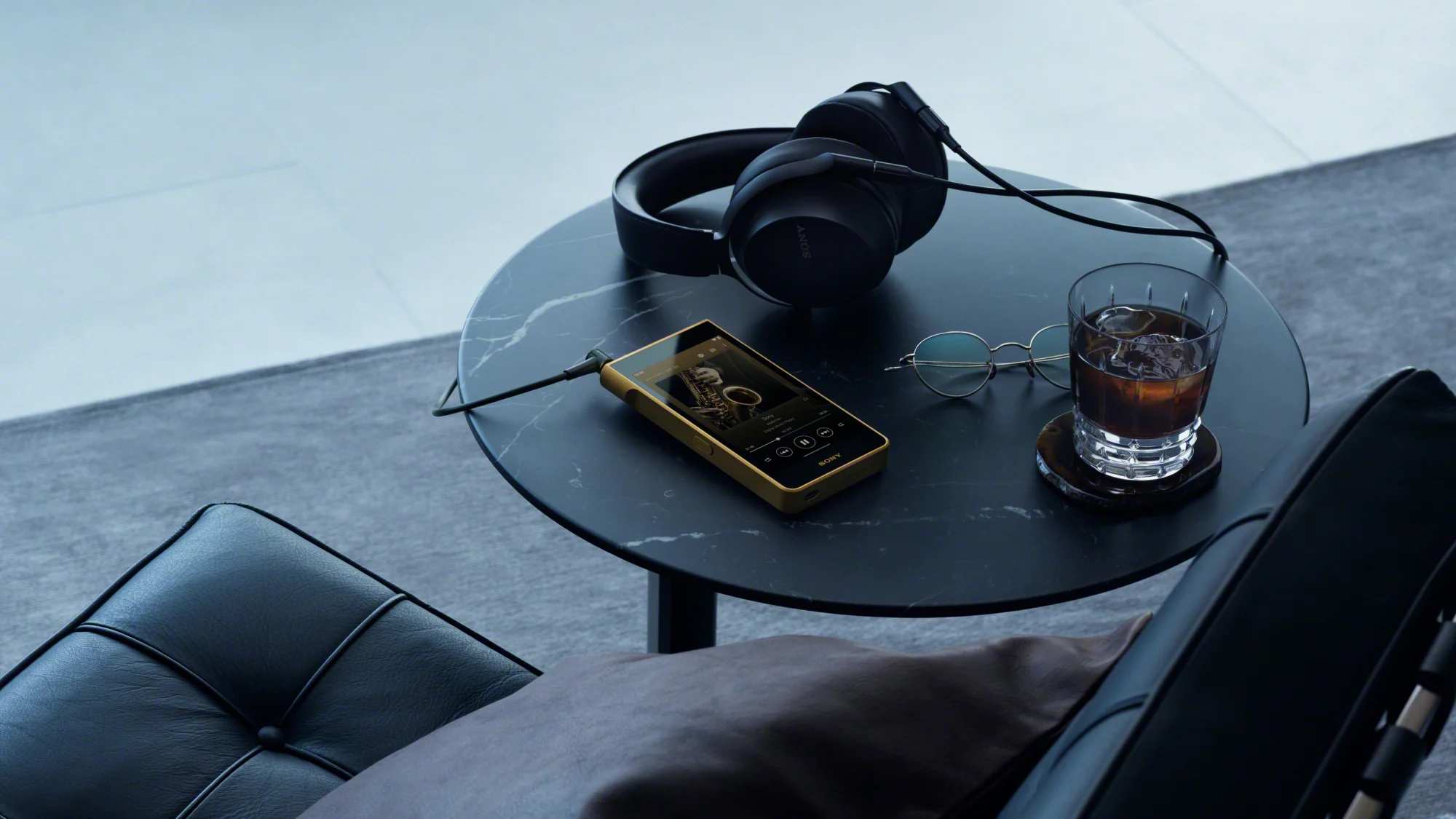 Un Walkman NW-WM1ZM3 doré, relié par un câble, posé sur une table ronde en marbre noir à côté d’un casque Sony noir, d’une paire de lunettes et d’un verre de boisson glacée, près d’un fauteuil en cuir noir.
