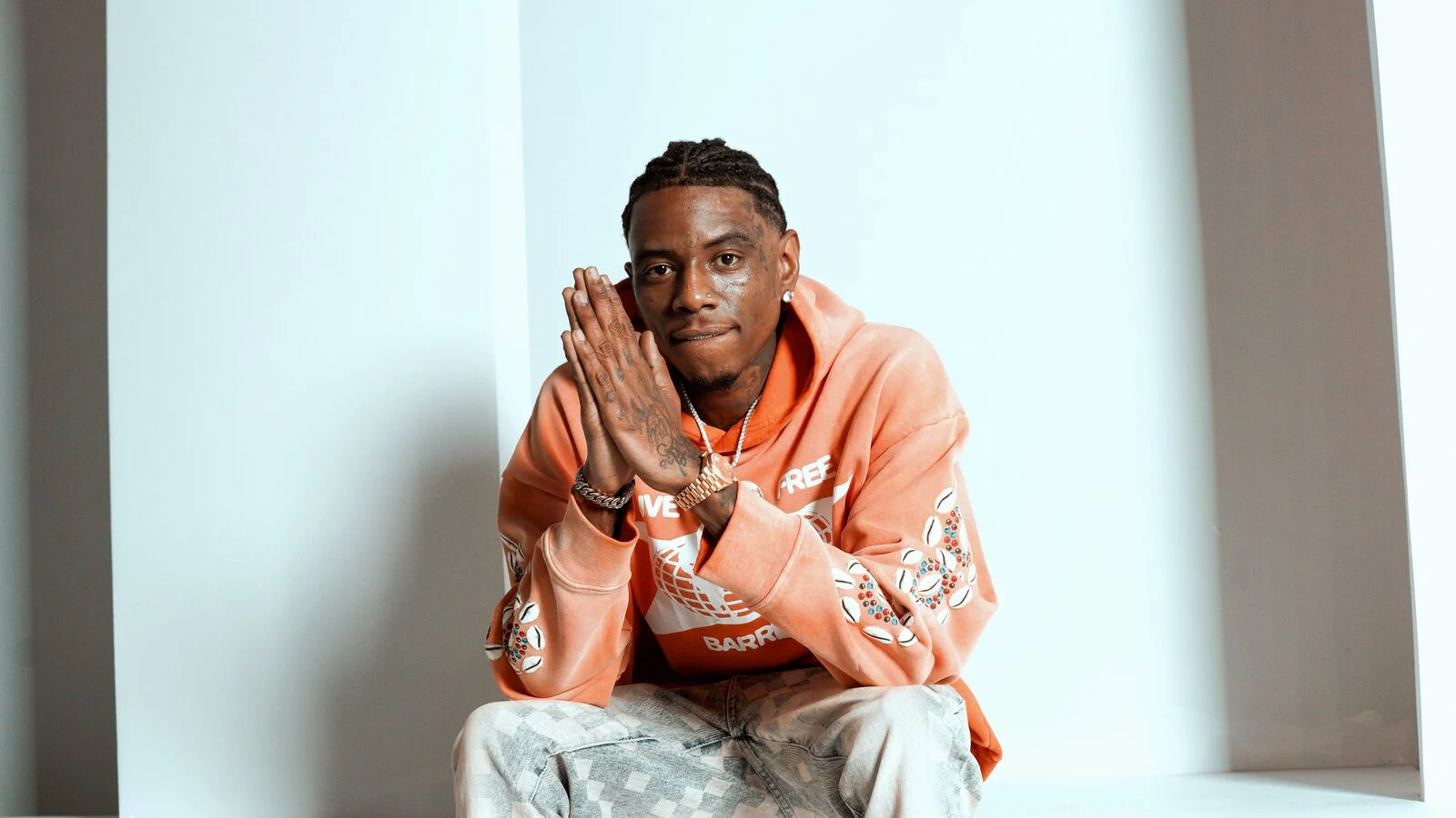 Soulja Boy, en sweat à capuche orange et jean délavé, assis sur des marches blanches, les mains jointes sous le menton, regarde l’objectif.