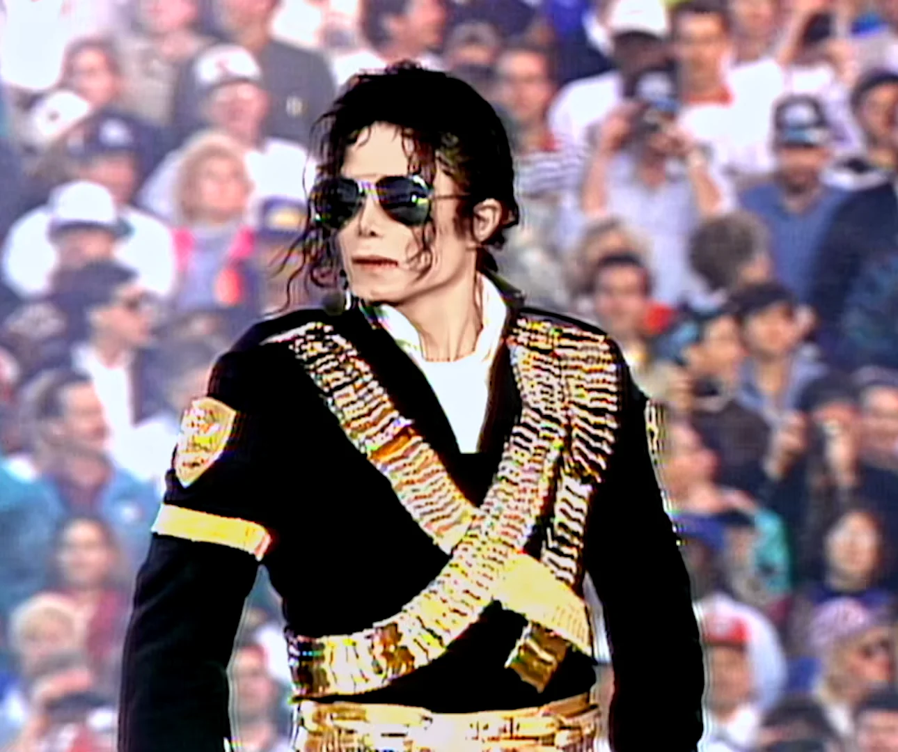 Michael Jackson sur la scène du Rose Bowl de Pasadena, le 31 janvier 1993, lunettes noires, veste noire de style militaire barrée de cartouchières dorées, devant une tribune pleine de spectateurs.