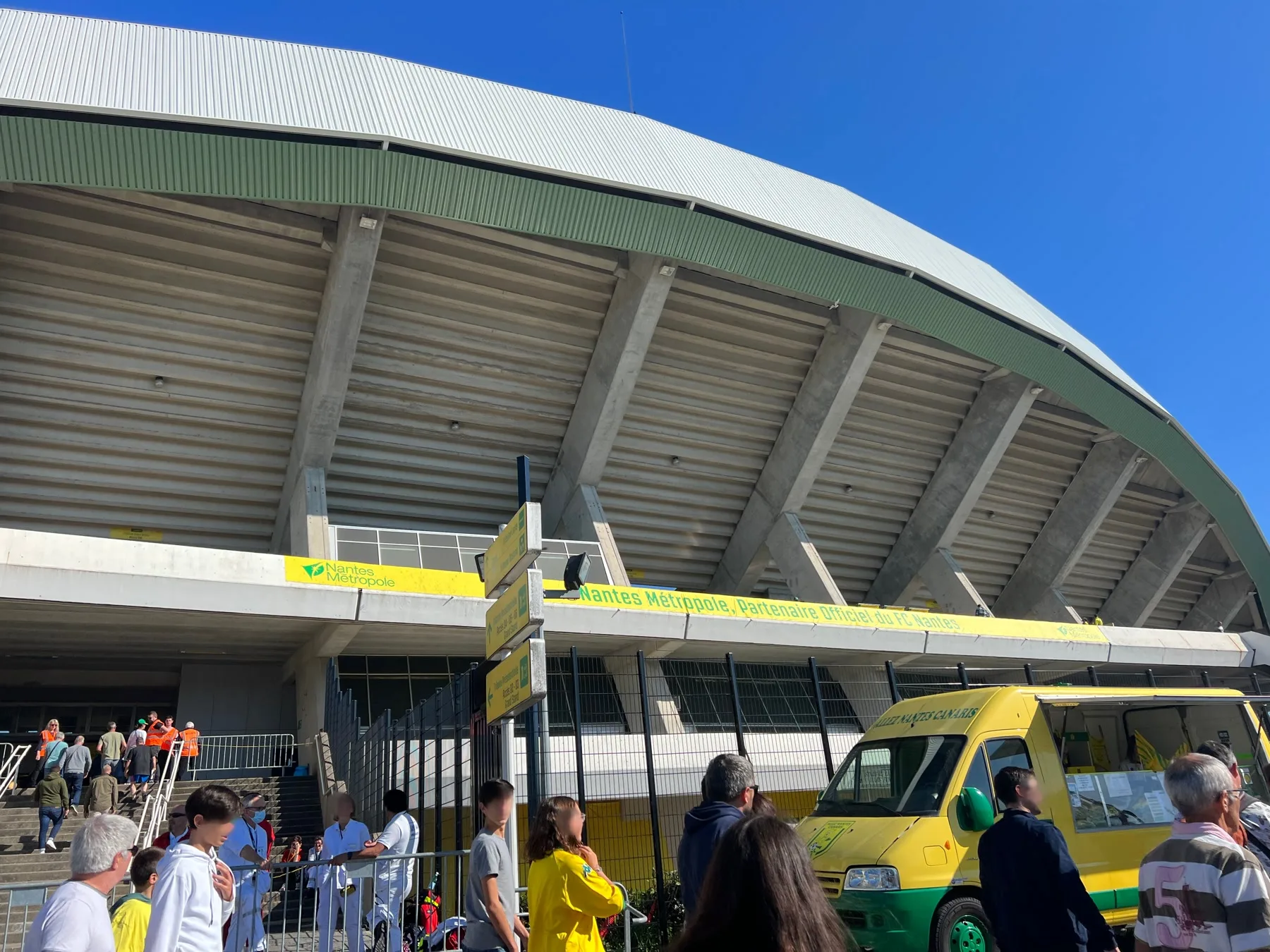 La tribune extérieure du stade de la Beaujoire à Nantes, sous un ciel bleu, avec des supporters devant l’entrée.