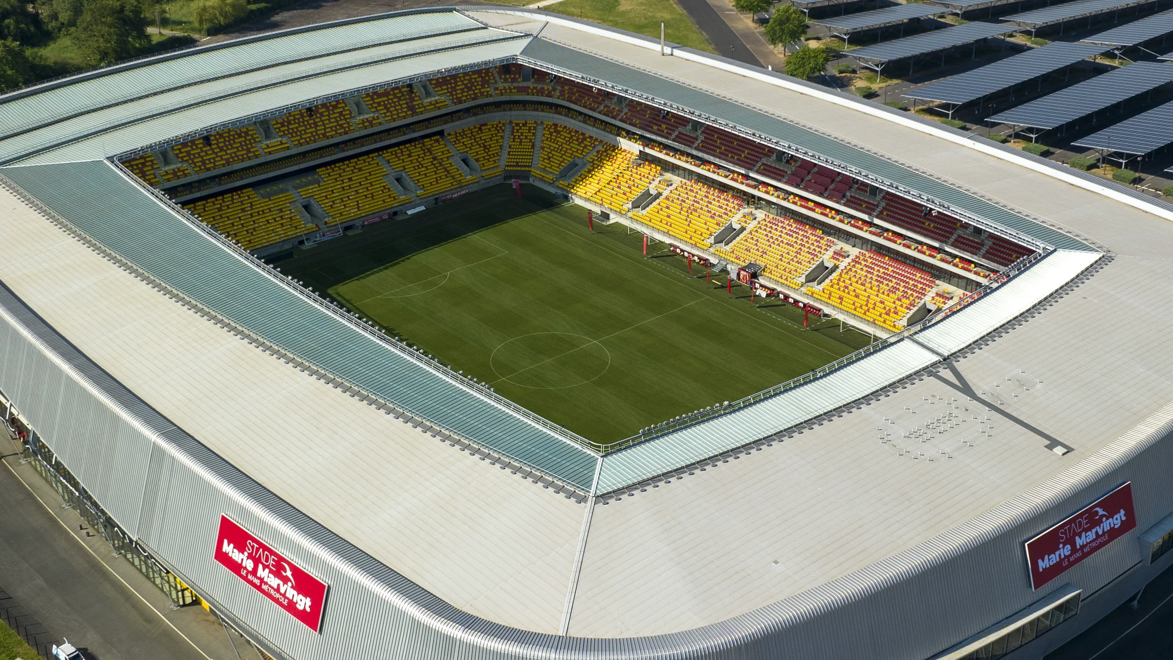 Vue aérienne du stade Marie-Marvingt du Mans par beau temps : l’enceinte au toit gris clair, ses tribunes rouge et or vides autour de la pelouse, et les ombrières photovoltaïques du parking à l’arrière-plan.
