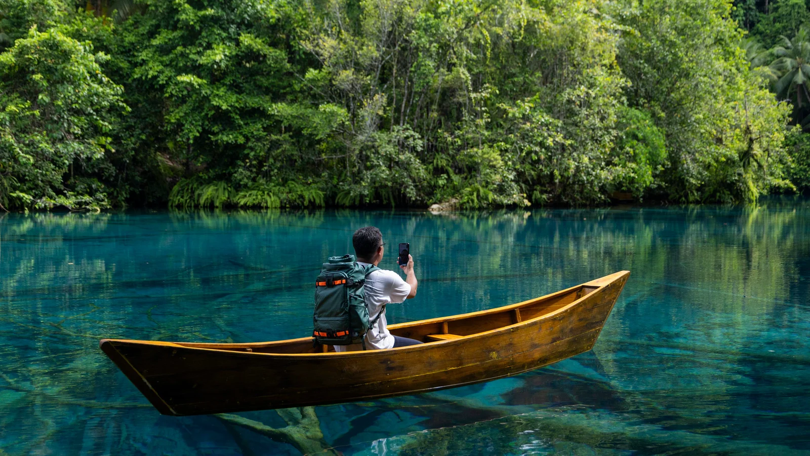 Un homme assis dans une pirogue en bois, sac à dos sur les épaules, lève son téléphone au milieu d’un lac turquoise bordé d’une forêt tropicale, loin de toute antenne.