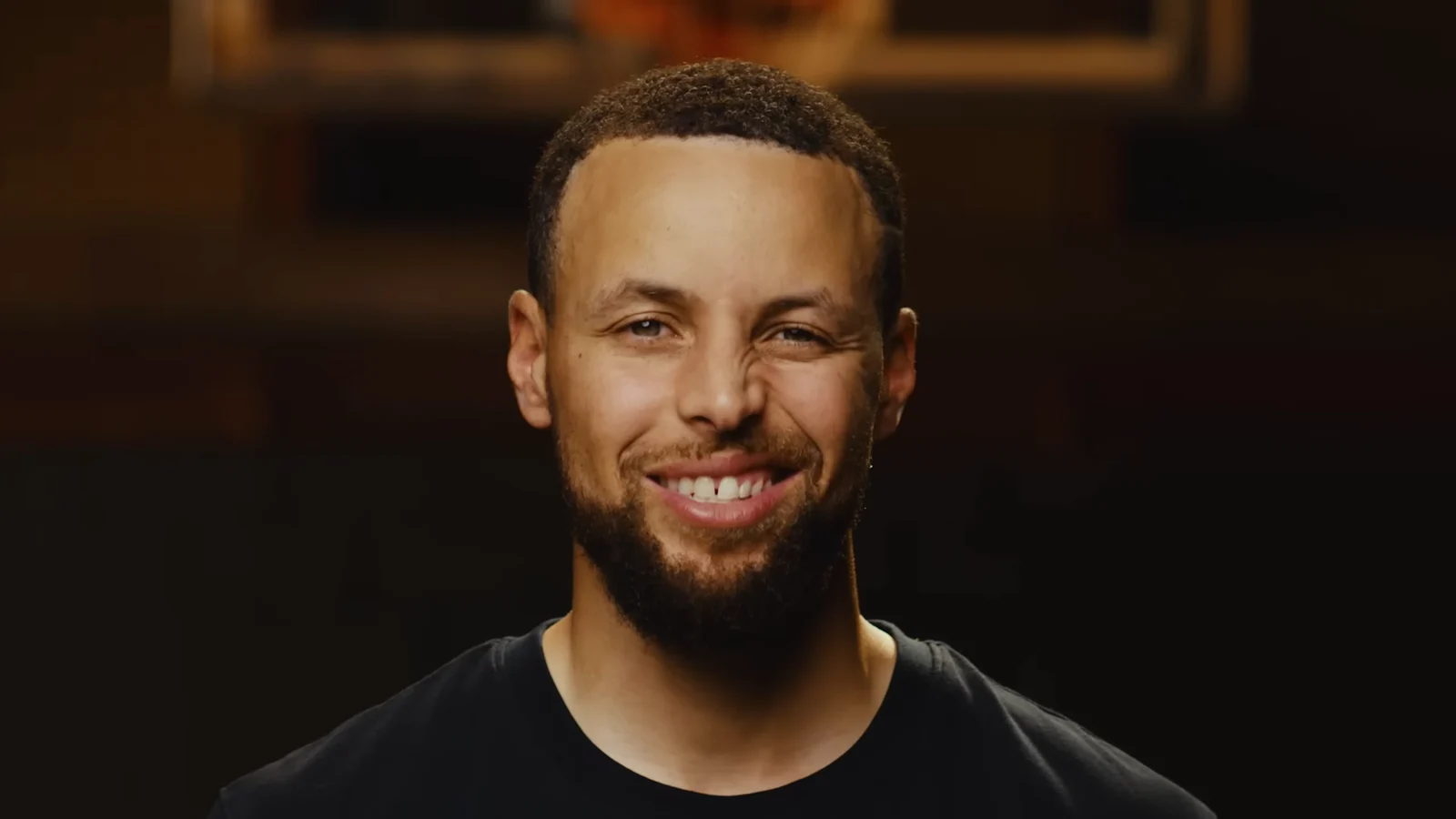 Stephen Curry, barbe courte et tee-shirt noir, sourit face caméra dans un gymnase plongé dans la pénombre ; un panier de basket se devine flou au-dessus de lui.