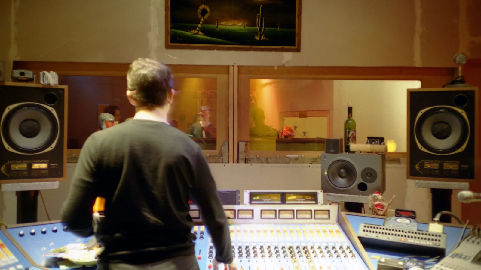 Un ingénieur du son, de dos, devant une grande console de mixage dans la cabine de contrôle d'un studio d'enregistrement, encadré par deux enceintes de monitoring, face à la vitre donnant sur la salle de prise