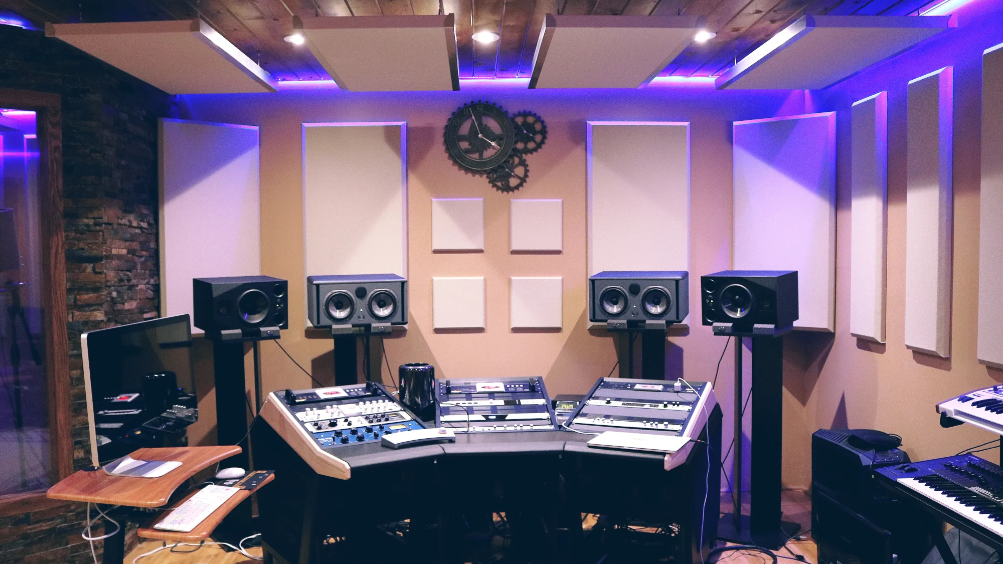 Un studio d’enregistrement éclairé en violet : poste de travail avec écran d’ordinateur, quatre enceintes de monitoring sur pieds, racks d’effets, claviers sur la droite et panneaux acoustiques aux murs.