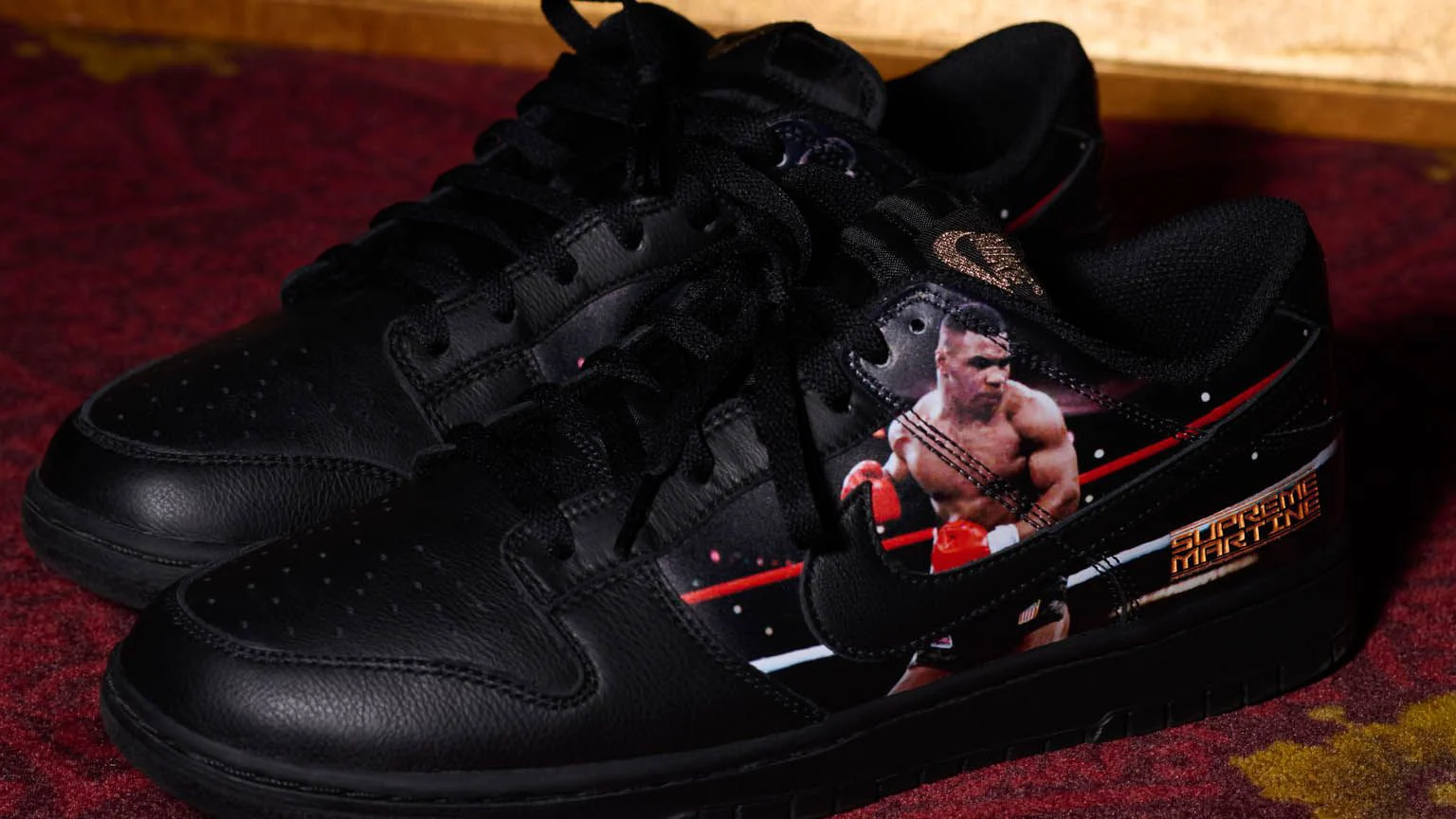 Une paire de Nike Dunk Low noires posée sur une moquette rouge devant des moulures dorées ; sur le flanc, une photo de Mike Tyson en gants rouges sur un ring et un lettrage doré « Supreme Martine ».