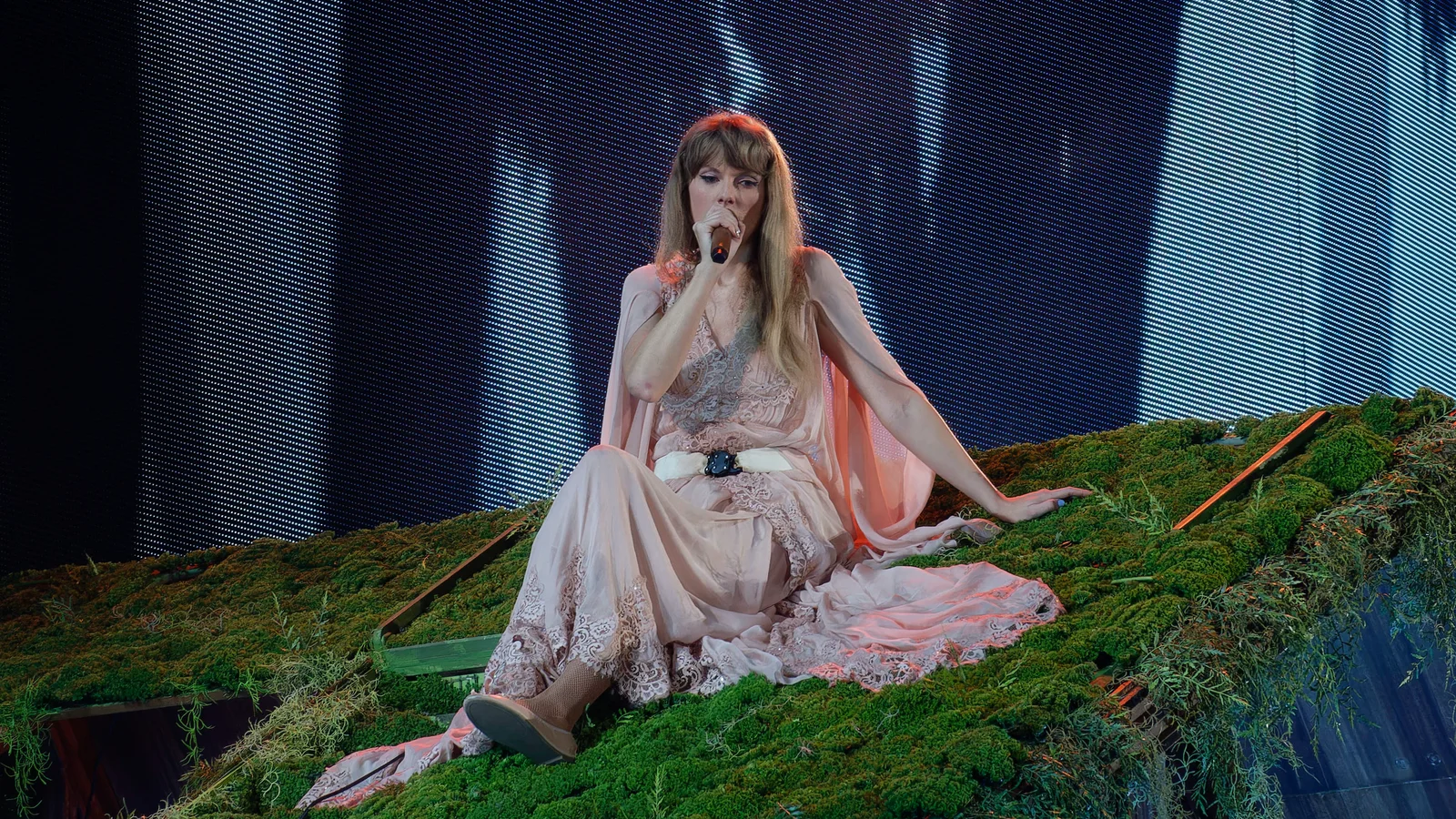 Taylor Swift assise sur un décor de mousse verte, en robe longue rose pâle à dentelle, chantant dans un micro devant un écran géant, lors de l’Eras Tour à Arlington, au Texas.