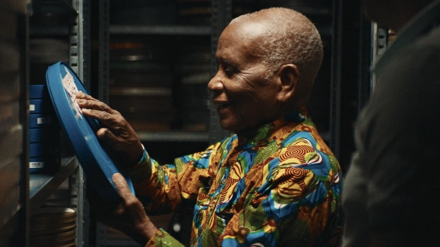 Chris Hesse, chemise imprimée aux motifs colorés, sourit en tenant une boîte de bobine bleue devant des rayonnages d’archives.