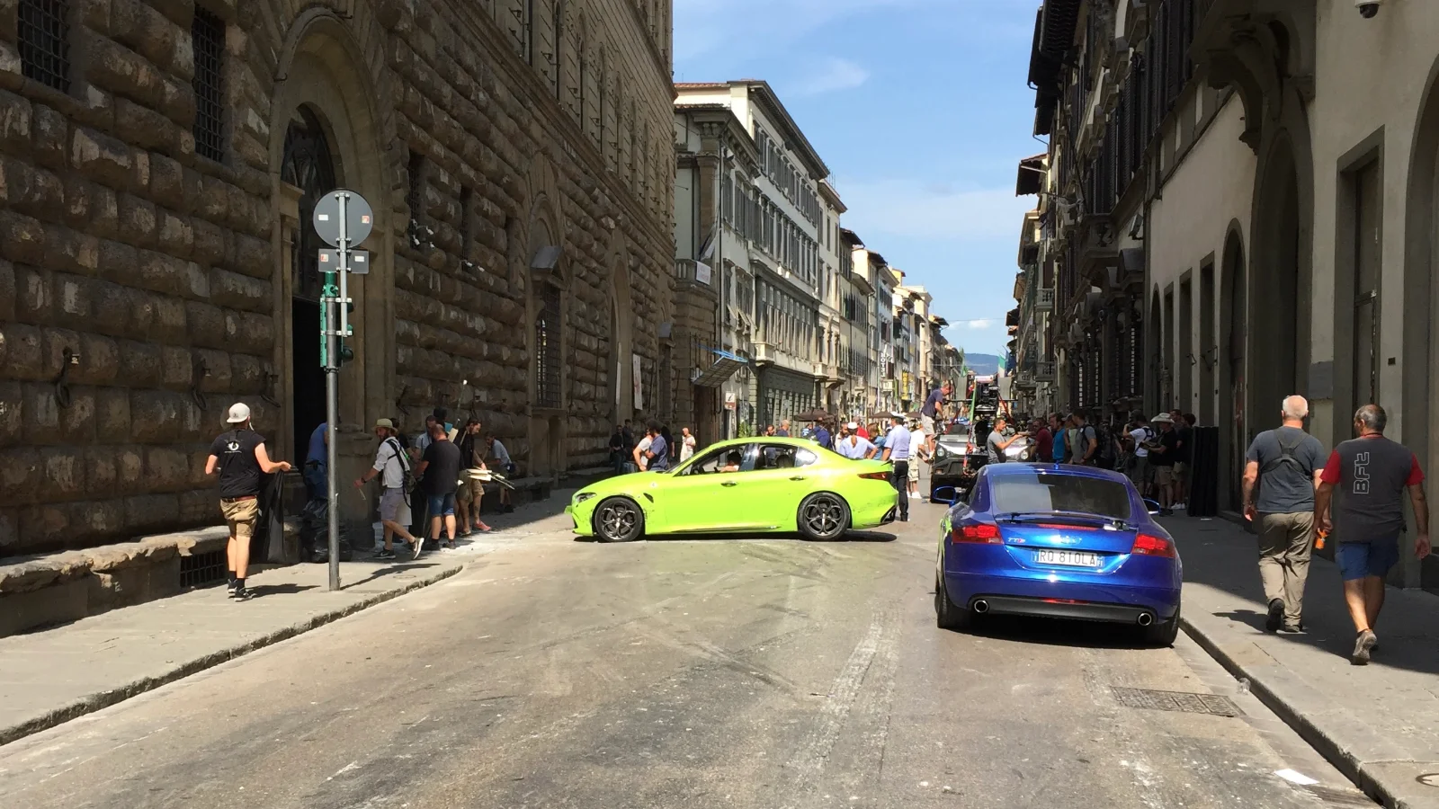 Rue de Florence bordée de palais en pierre pendant un tournage : une Alfa Romeo Giulia vert vif est arrêtée en travers de la chaussée, entourée de techniciens et de badauds, tandis qu'une voiture de sport bleue est garée au premier plan