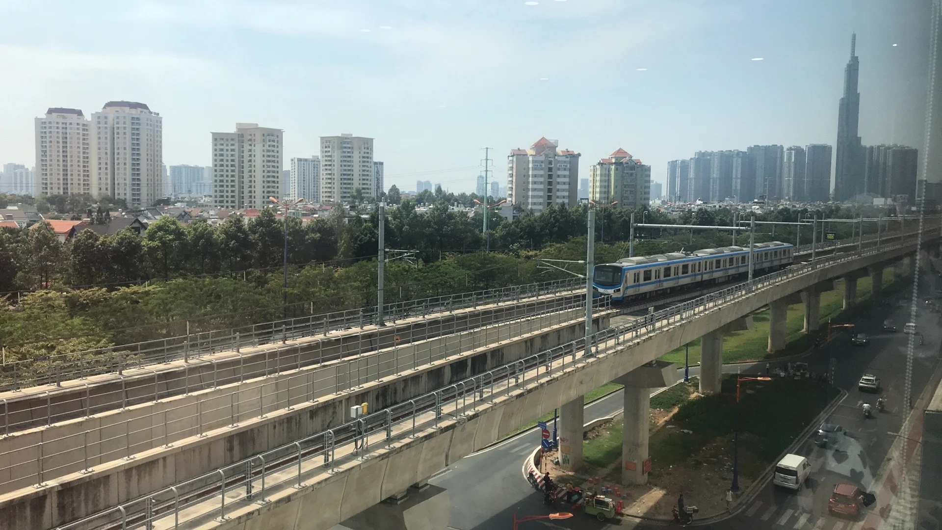 Une rame blanche et bleue de la ligne 1 du métro d’Hô Chi Minh-Ville circule sur un viaduc, avec des immeubles et la tour Landmark 81 au loin.