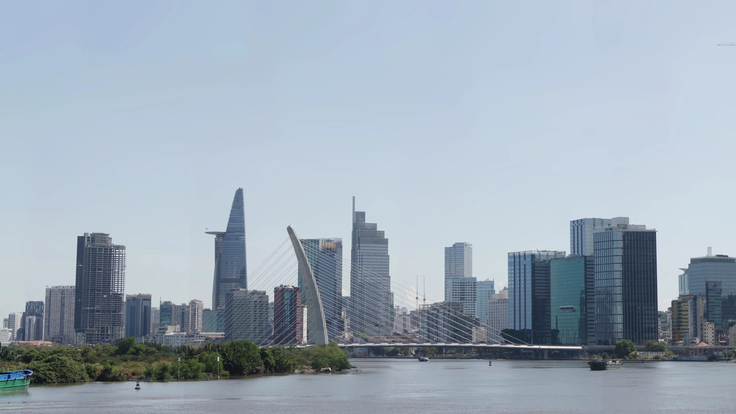 Les tours du centre d’Hô Chi Minh-Ville au bord de la rivière Saïgon, avec la tour Bitexco en forme de lotus et le pont à haubans de Thủ Thiêm, sous un ciel clair.