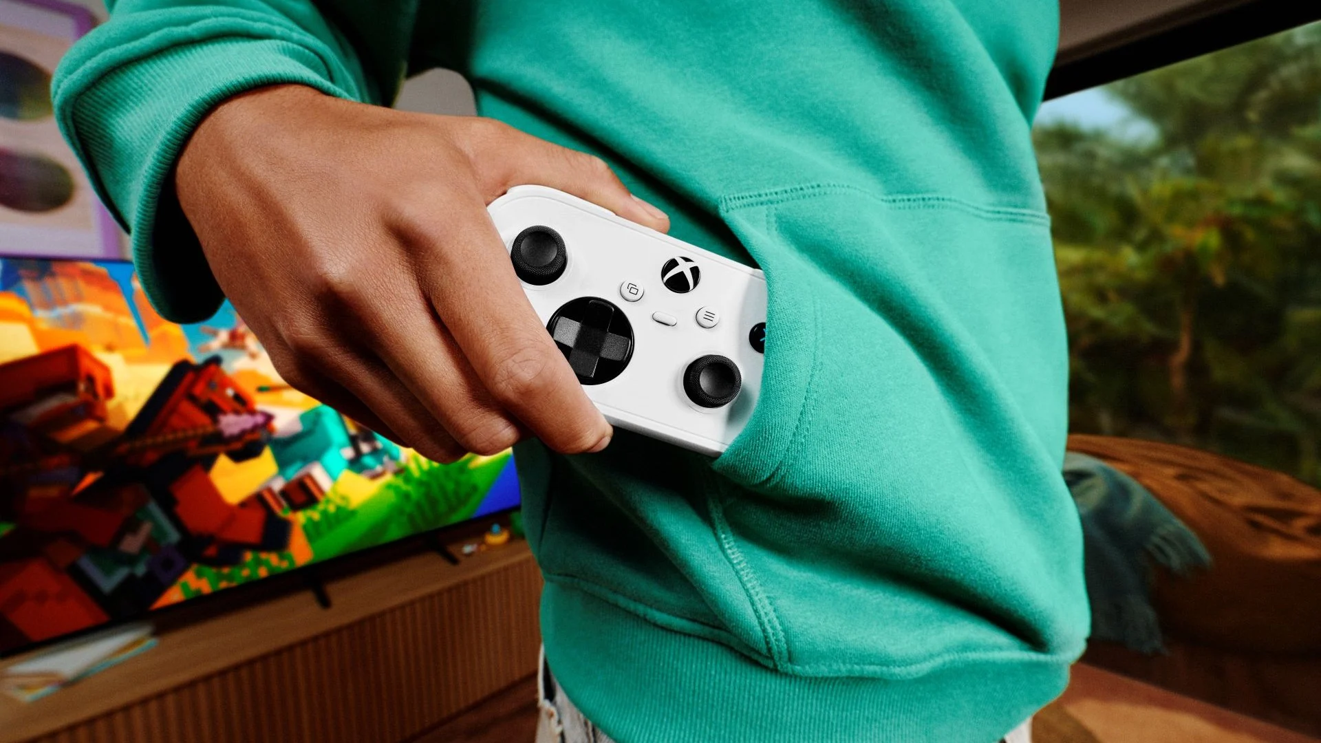 Une main sort une petite manette Xbox Nano blanche de la poche ventrale d’un sweat à capuche vert, devant un téléviseur allumé sur un jeu aux graphismes en cubes.