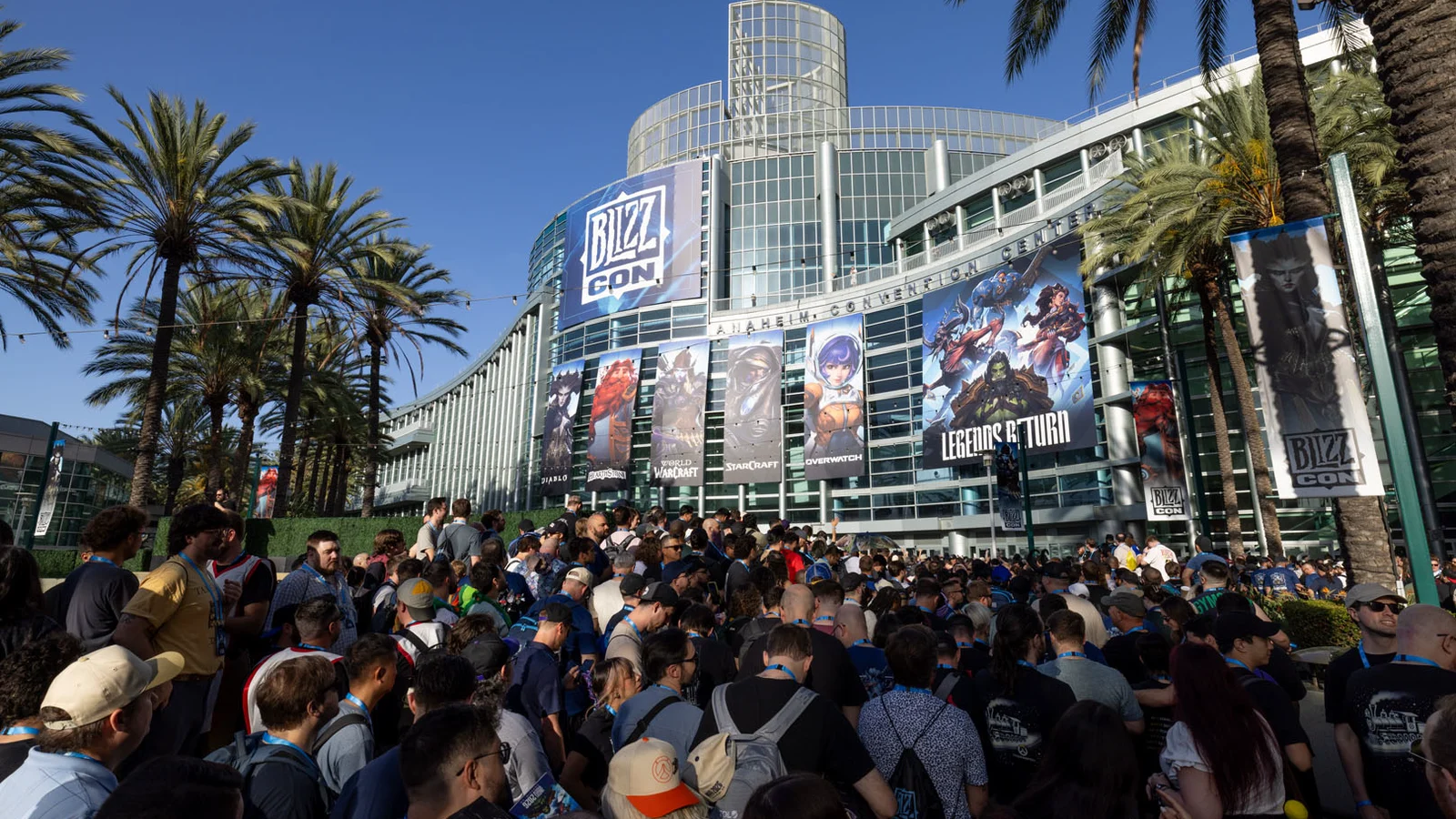 Une foule de visiteurs devant le centre de congrès d’Anaheim, sous les palmiers, avec de grandes bannières BlizzCon et des affiches de jeux Blizzard sur la façade vitrée.