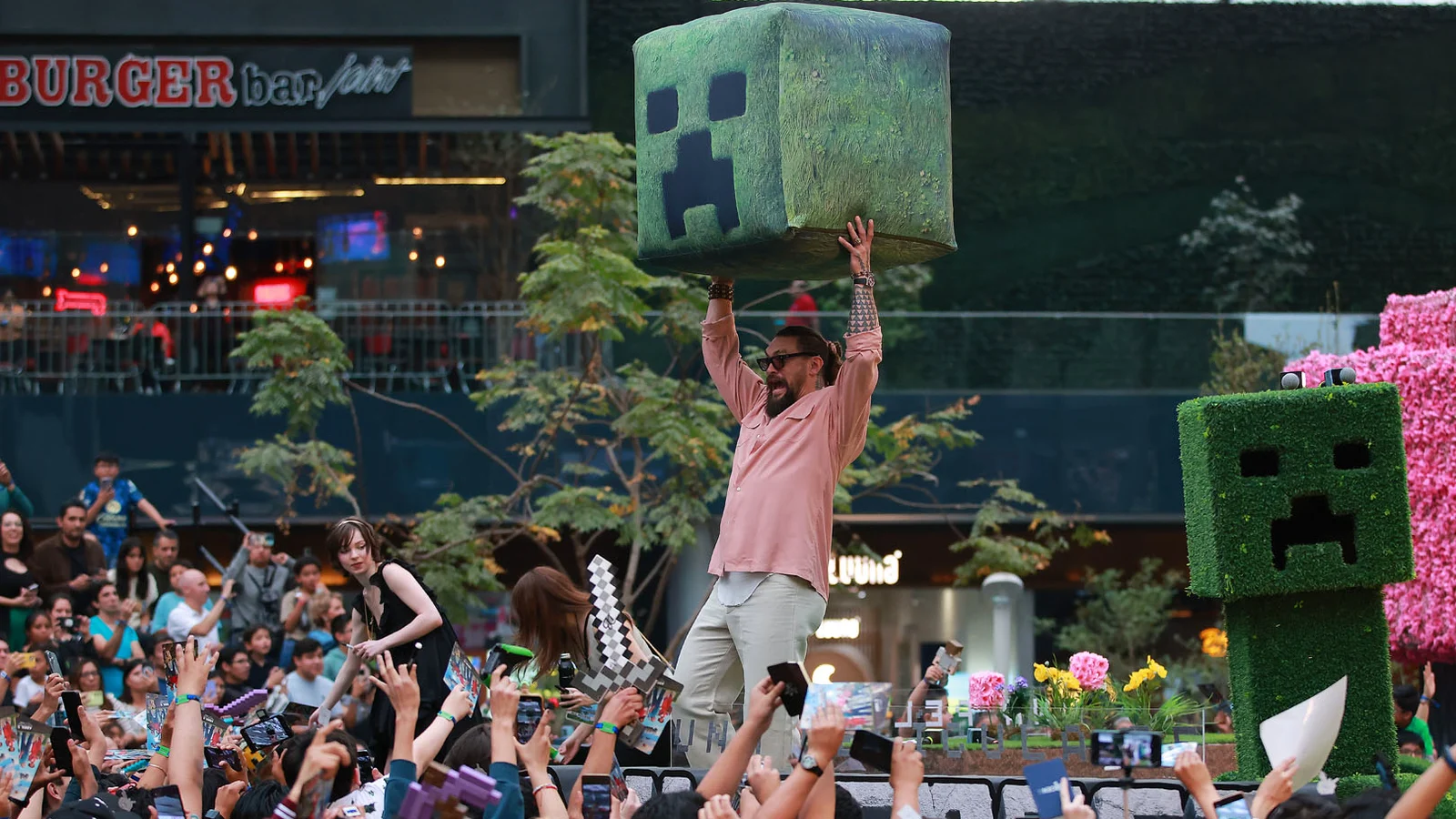 Jason Momoa, chemise rose, brandit à bout de bras une grosse tête de Creeper verte au-dessus d’une foule de fans qui le photographient, lors d’un événement autour du film Minecraft.