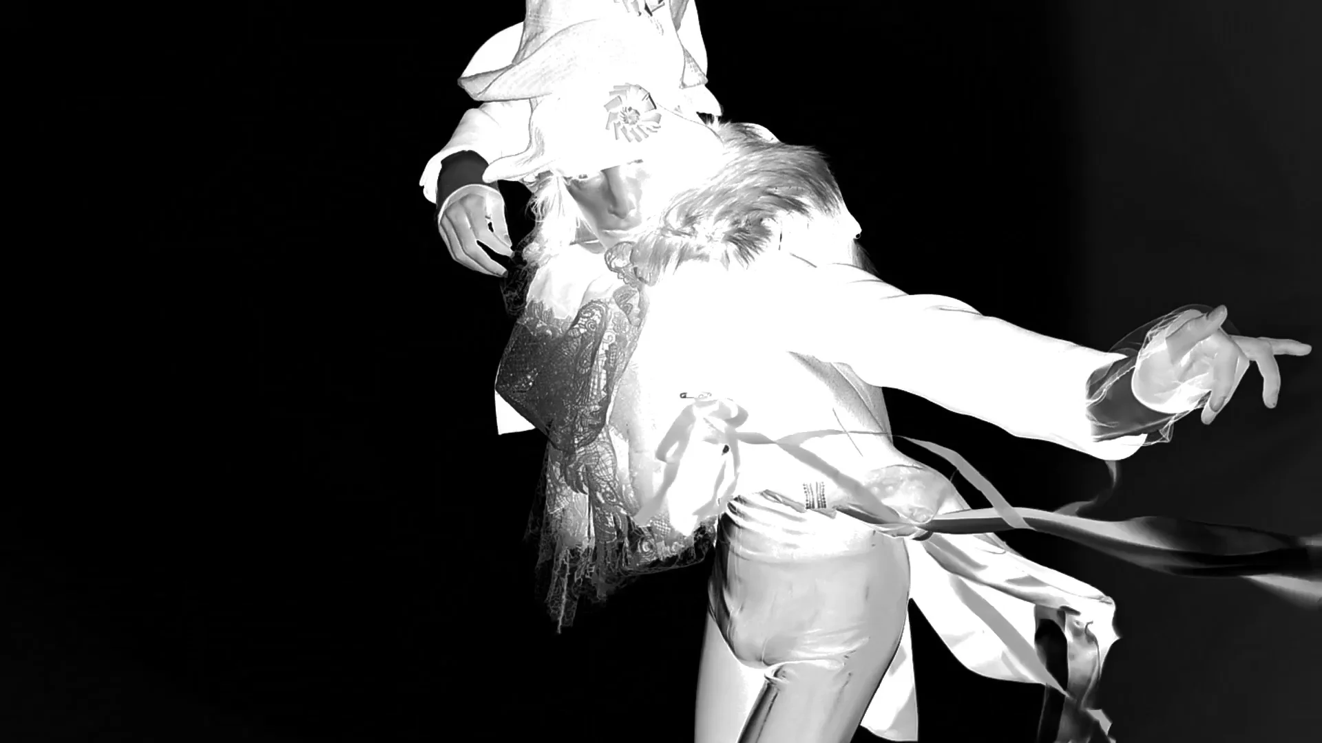 Image en noir et blanc aux tons inversés tirée de la vidéo officielle de Re{form} Zara : un mannequin en veste blanche à basques, col de fourrure et jabot de dentelle, bras tendu, des rubans flottant à la taille, sur fond noir.